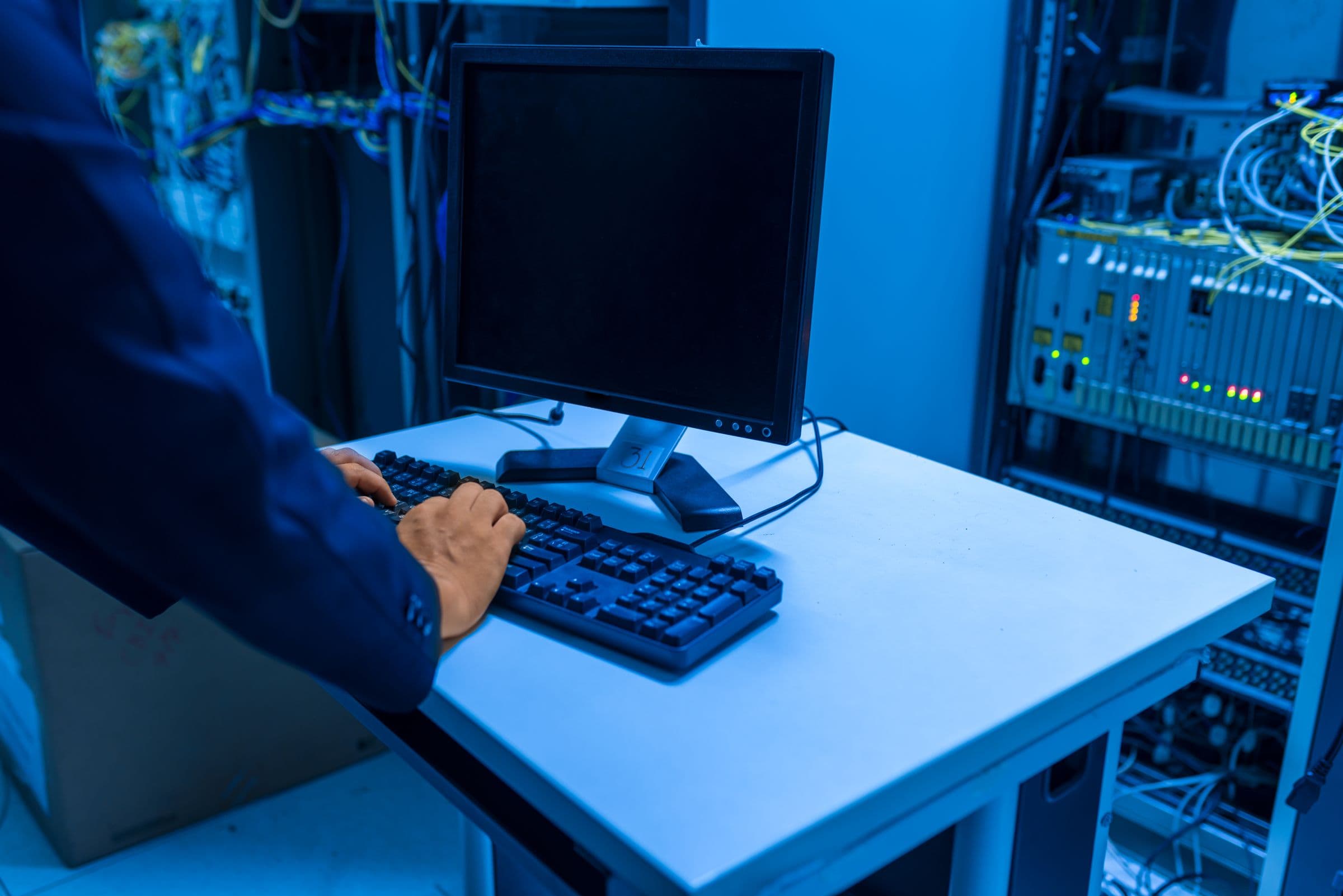 An IT technician at a console inside a blue-lit server room, hands on the keyboard, network equipment and patch leads racked behind.