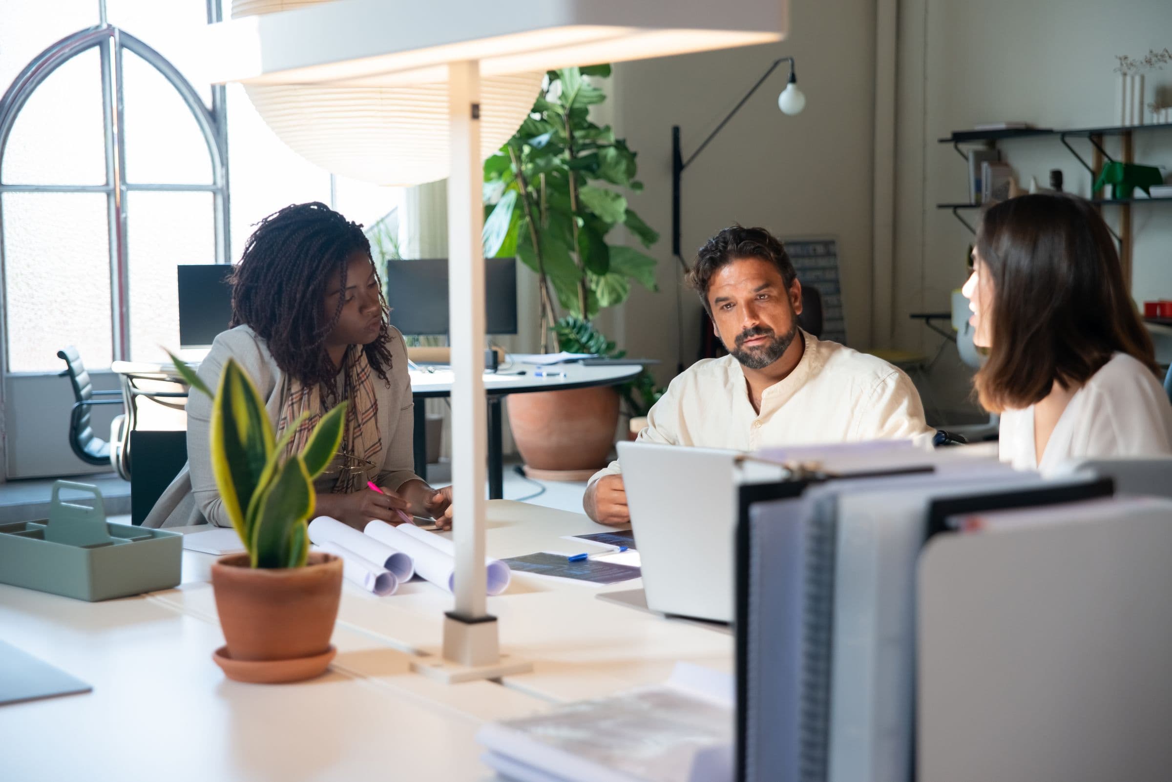 Three colleagues working together at a shared desk in a bright plant-filled studio.