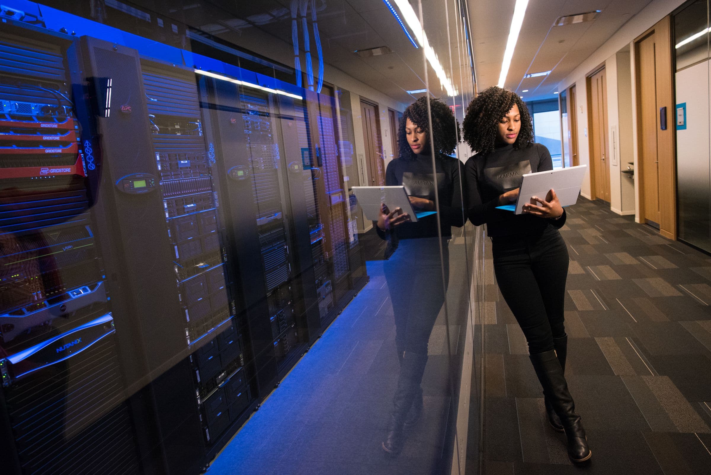 Two engineers walking a lit data-centre corridor, tablets in hand.
