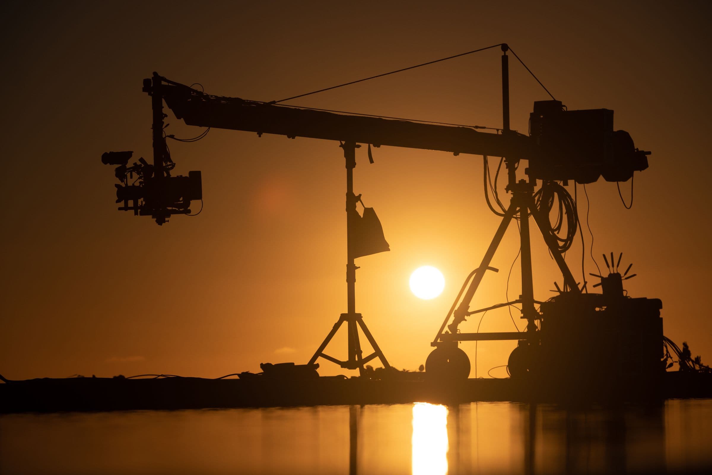 A camera crane silhouetted against the sun setting over open water.