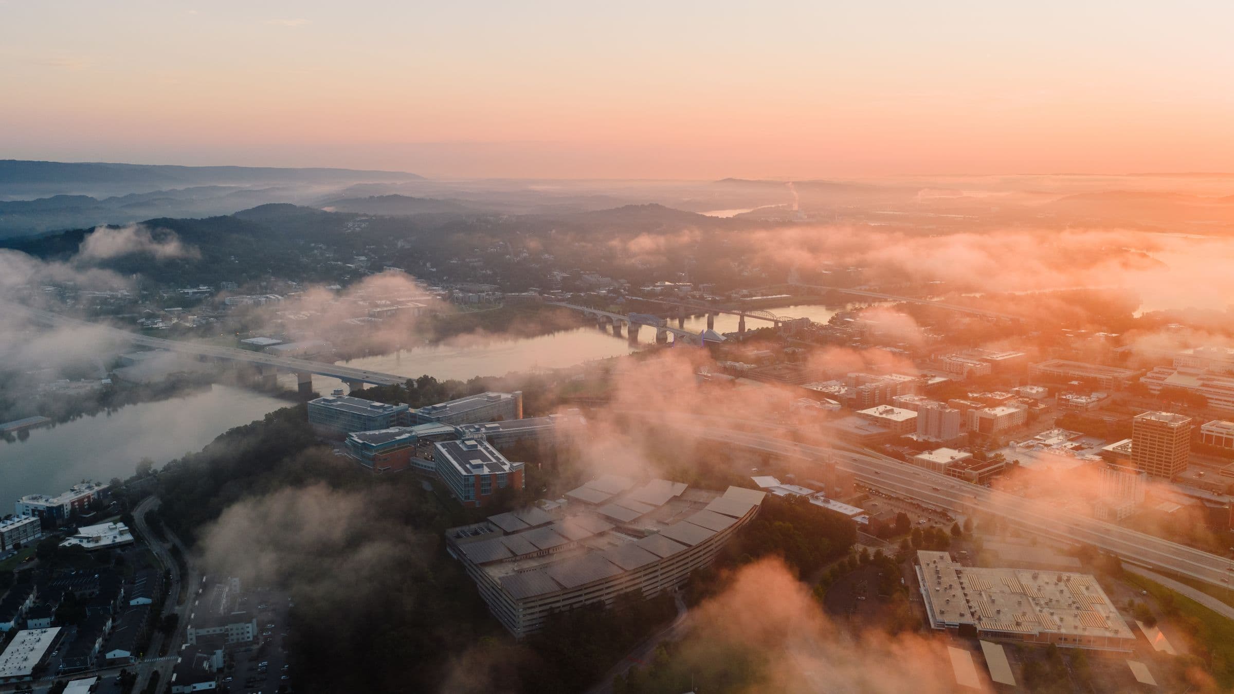 Low cloud drifting over a river city at sunrise, seen from the air.