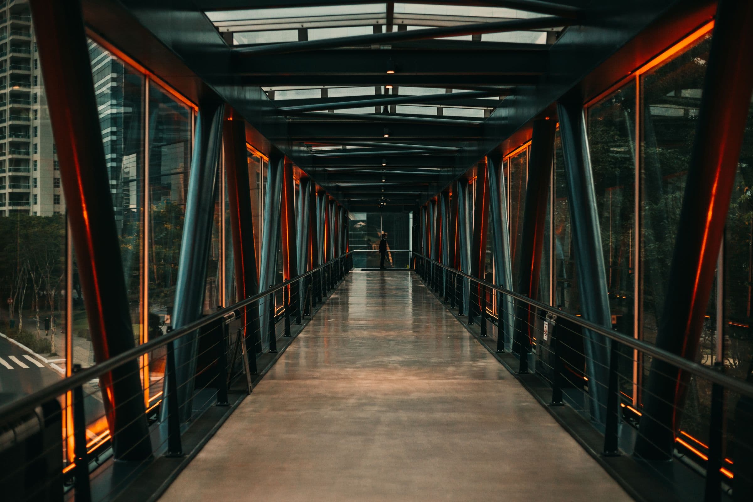 A glazed steel footbridge, its repeating trusses converging toward a distant opening.