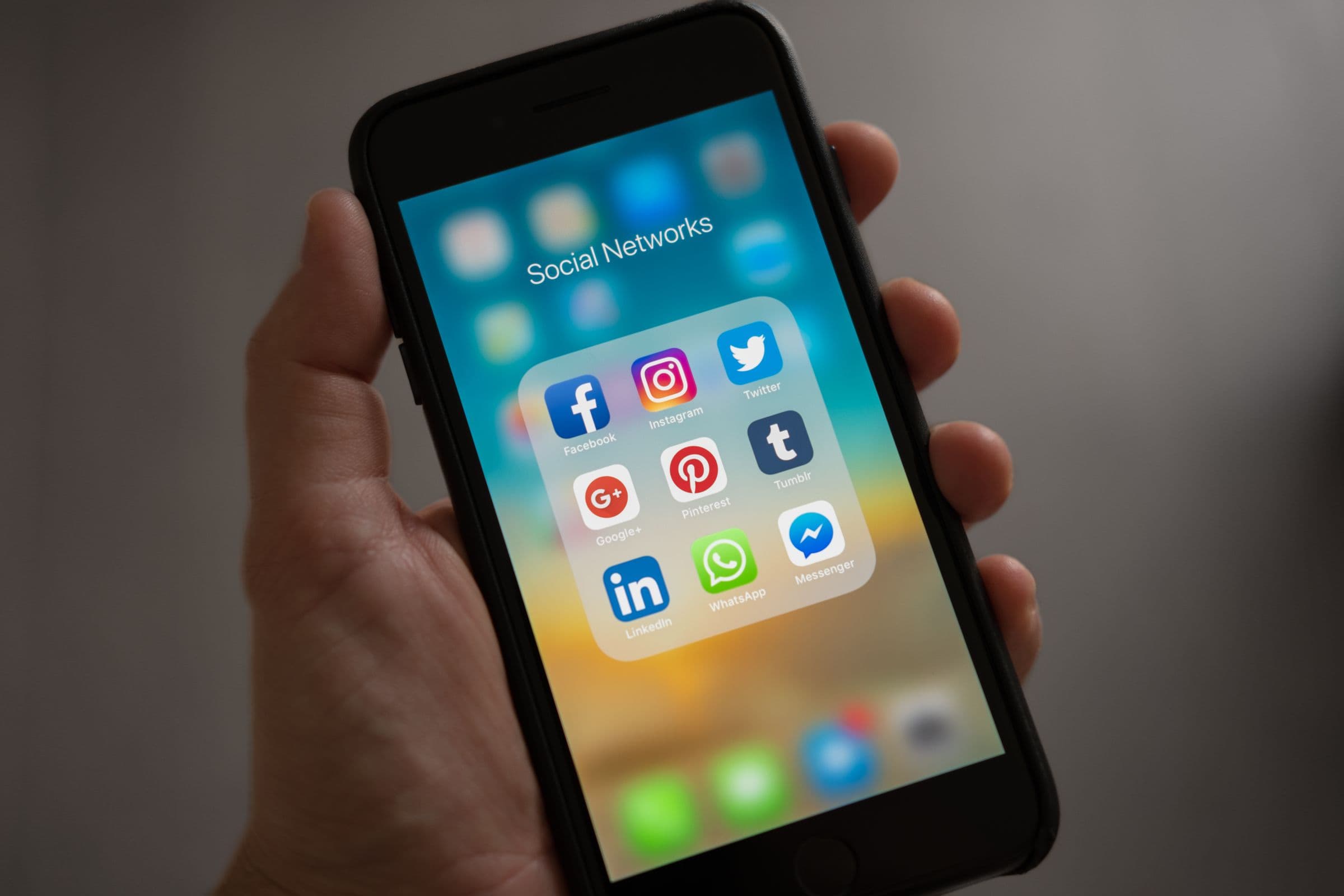 A hand holding a smartphone open on a folder of social network apps — Facebook, Instagram, Twitter, LinkedIn, WhatsApp and Messenger.