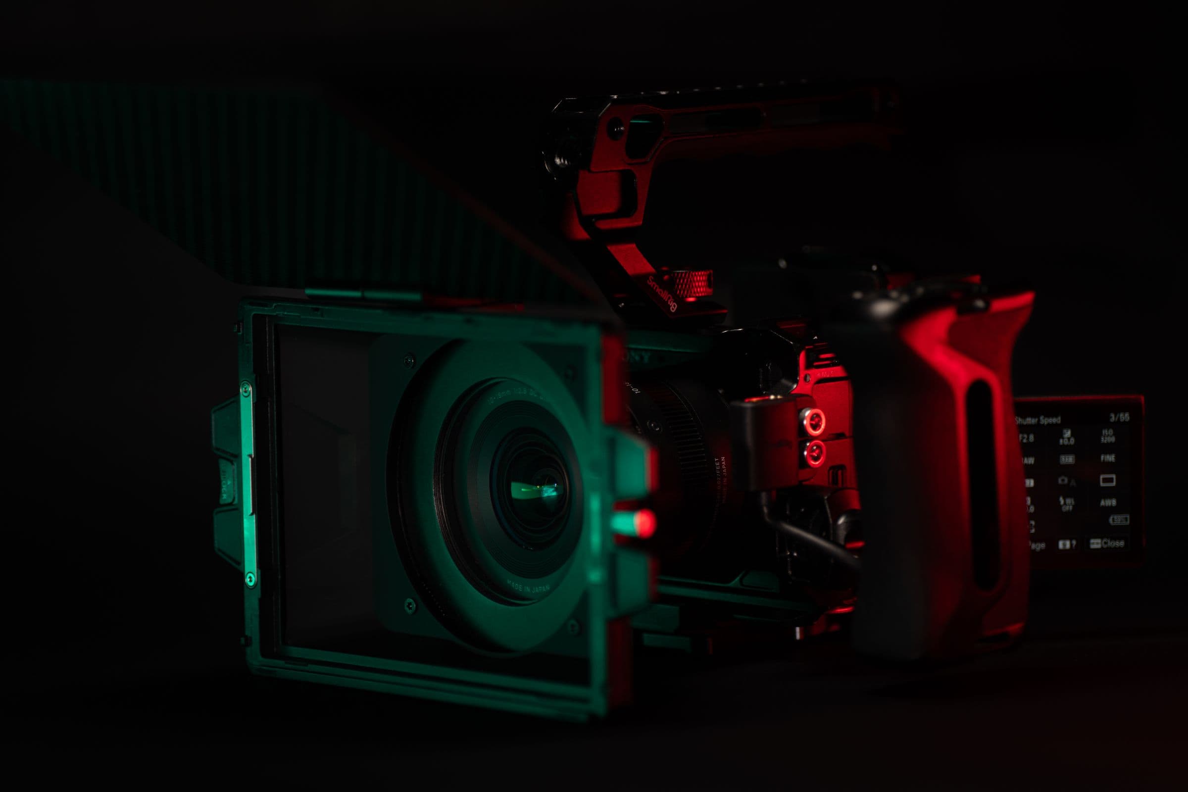 A cinema camera and matte box picked out by red and teal light in a dark studio.